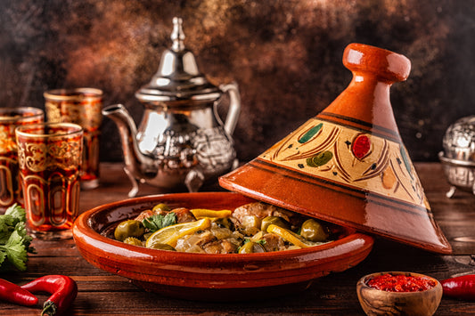Tajine de poulet aux olives et citrons confits dans un plat en terre cuite avec théière marocaine en arrière-plan.